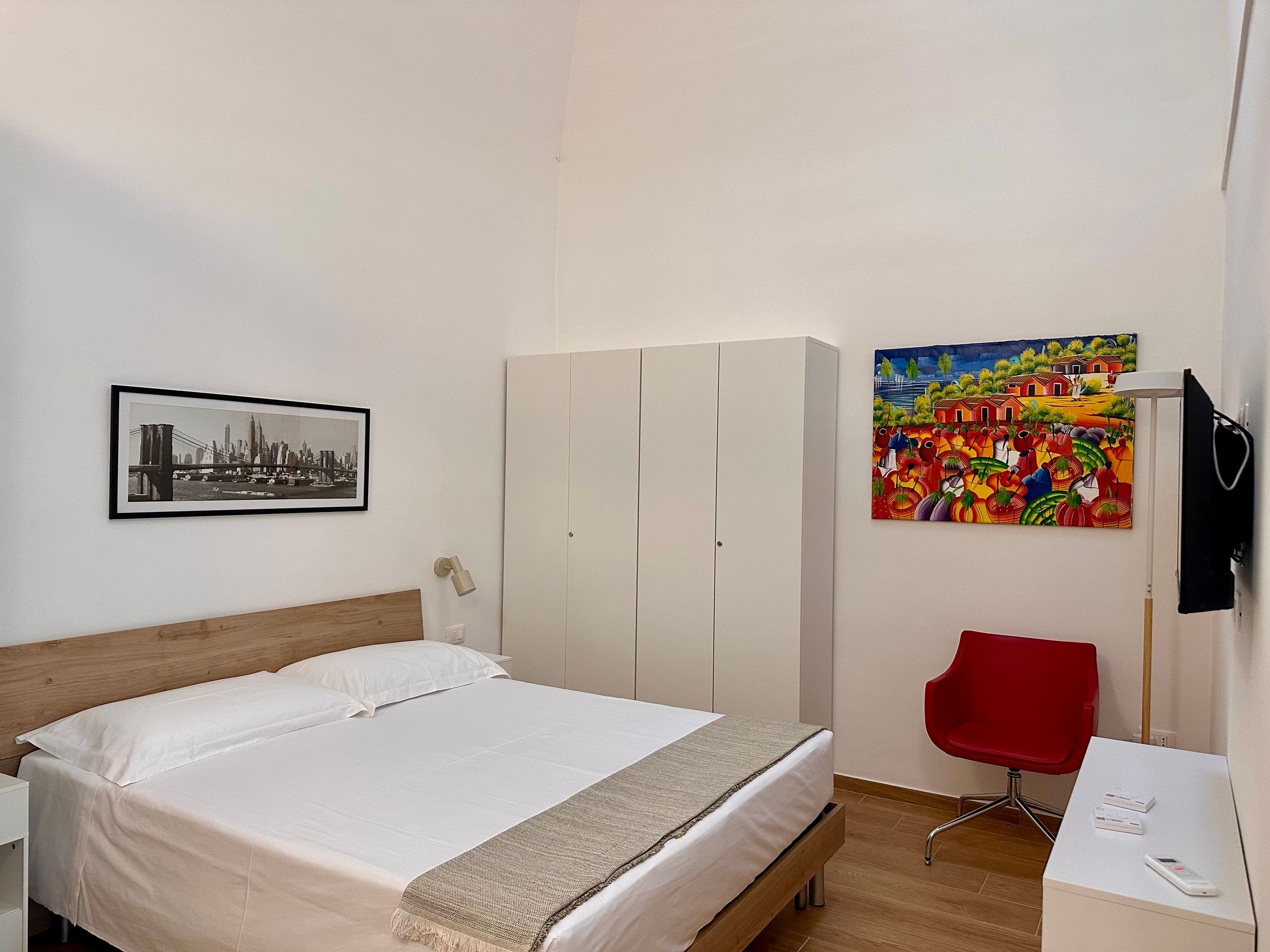 La camera matrimoniale con la testiera in legno, l’armadio bianco, la poltroncina rossa e un quadro colorato